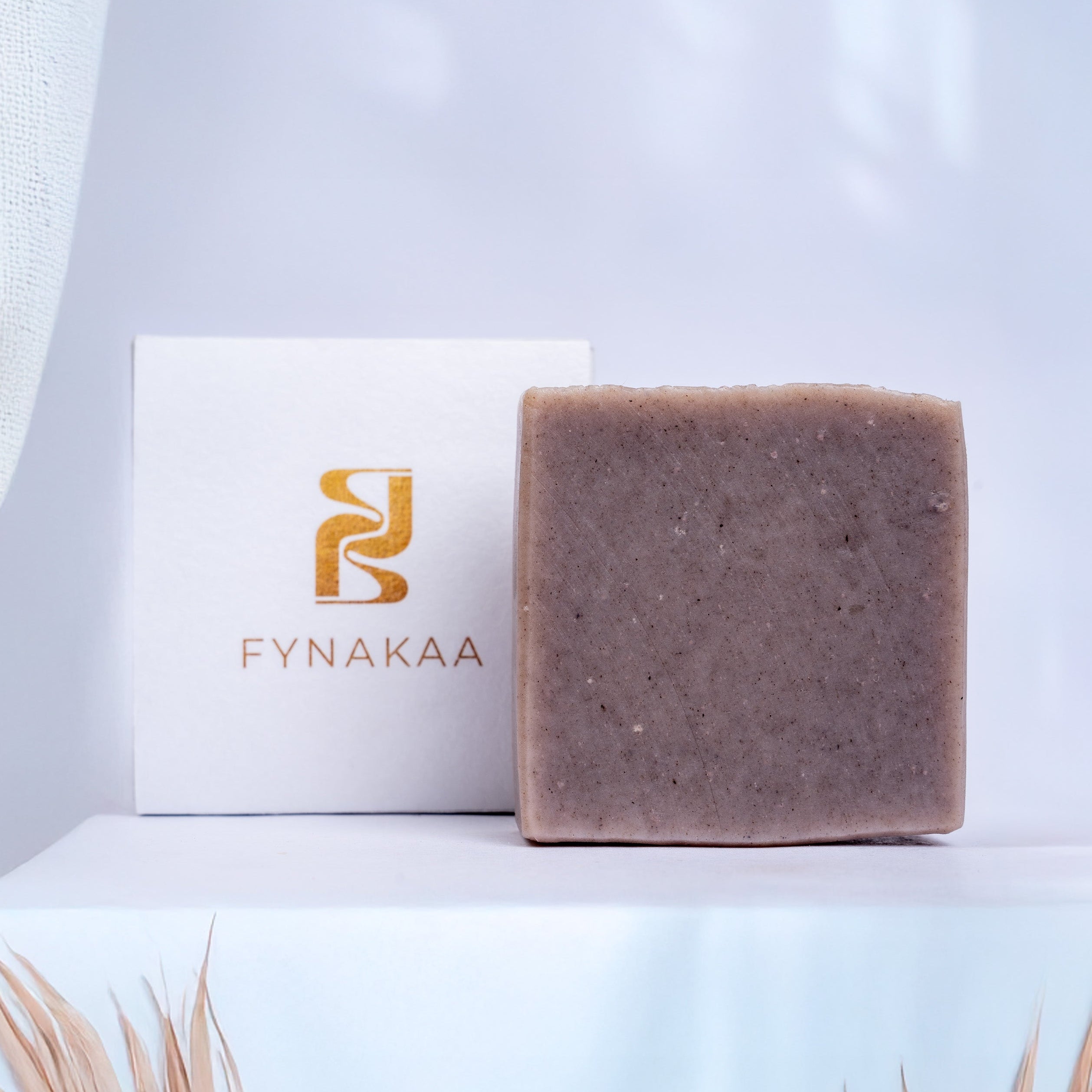 Bar of soap with a Fynakaa box on a light blue background with dried palm leaves.