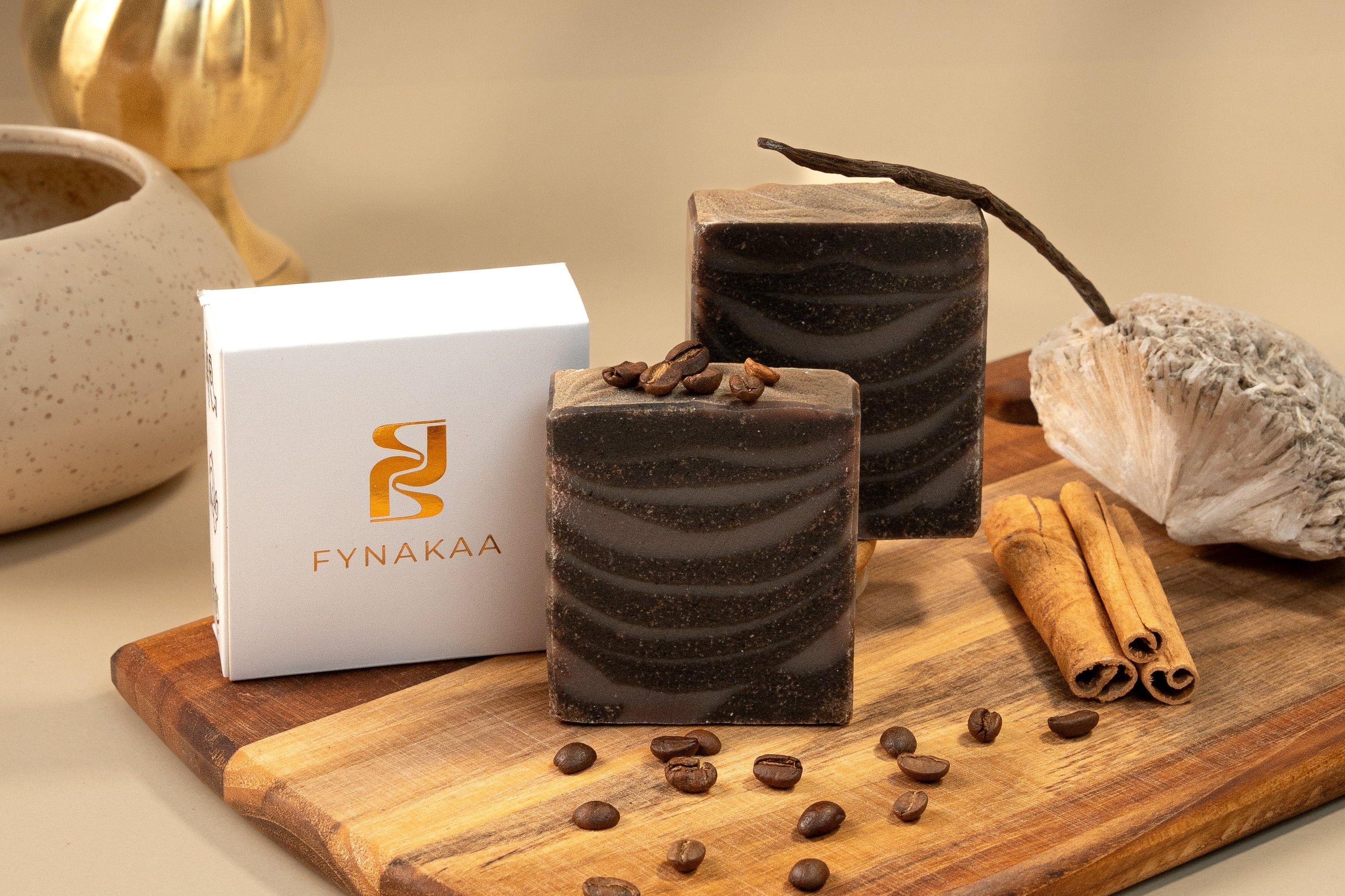 Two coffee soaps on a wooden board with coffee beans, cinnamon sticks, and a box labeled 'Fynakaa' against a beige background.