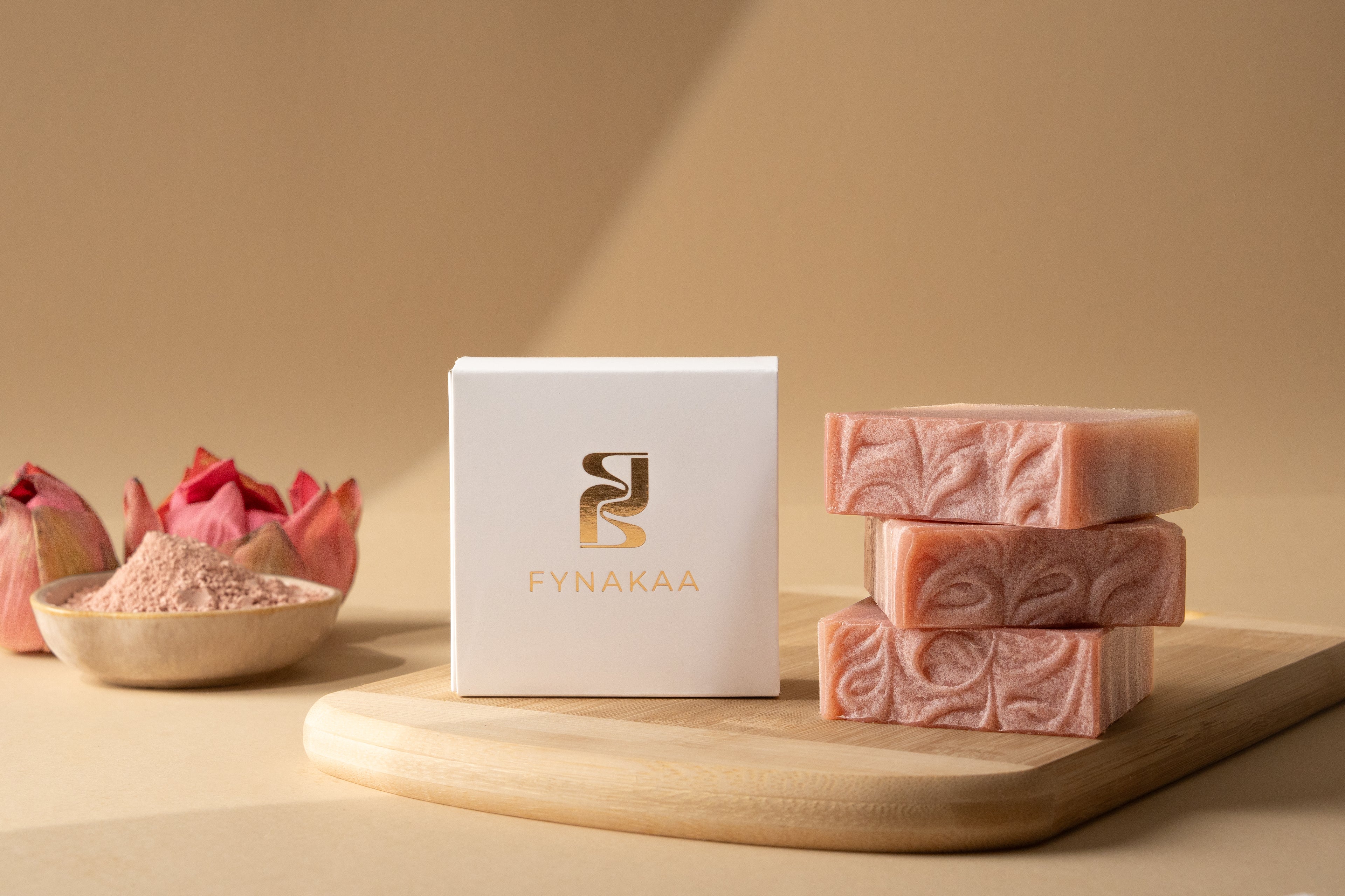 Pink soap bars and packaging with 'Fynakaa' brand on a wooden board against a beige background