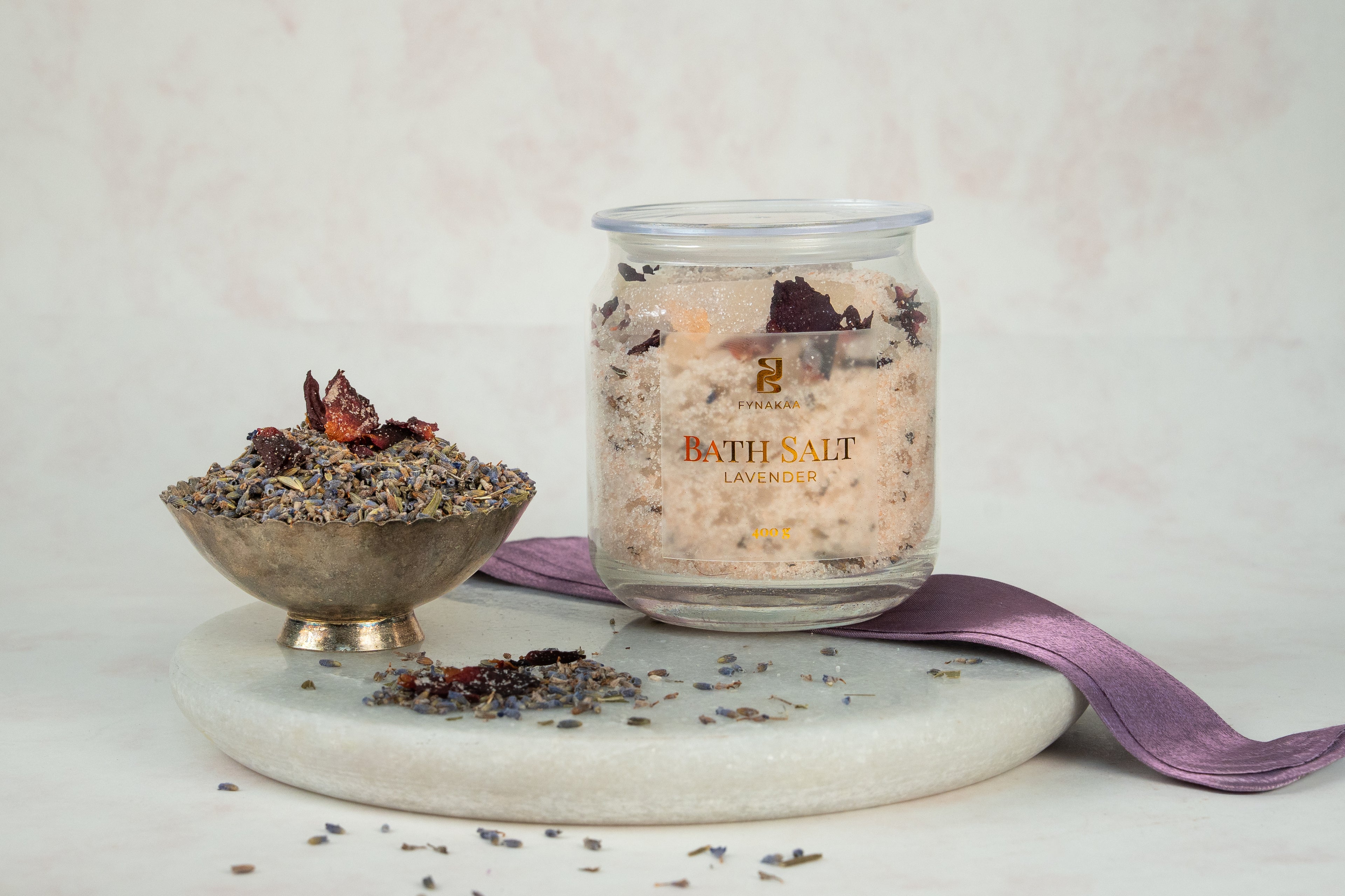 Jar of bath salt with a bowl of lavender buds and rose petals on a marble tray with a purple ribbon.