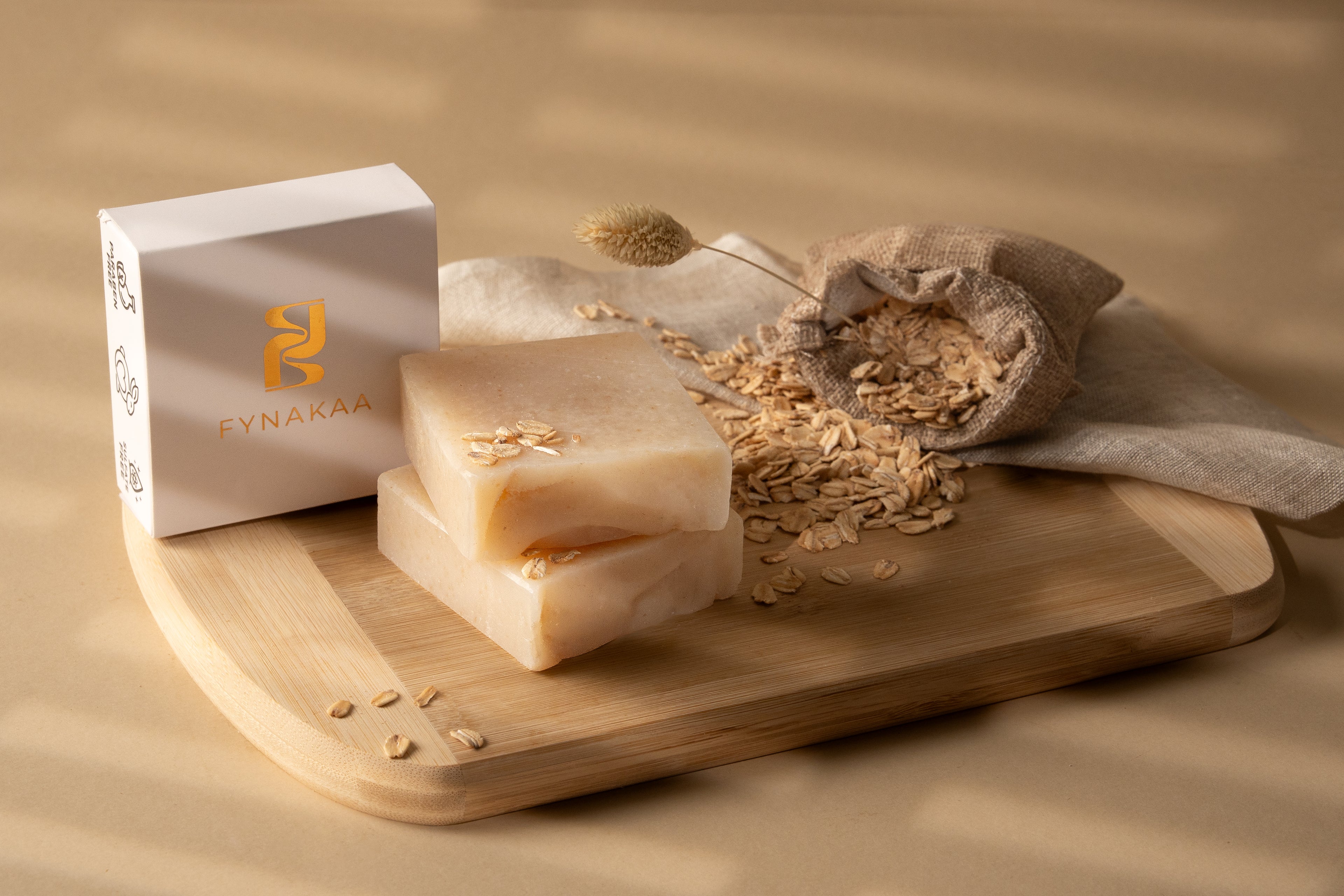 Two bars of soap with oatmeal on a wooden board next to a box labeled 'Fynakaa' on a beige background.