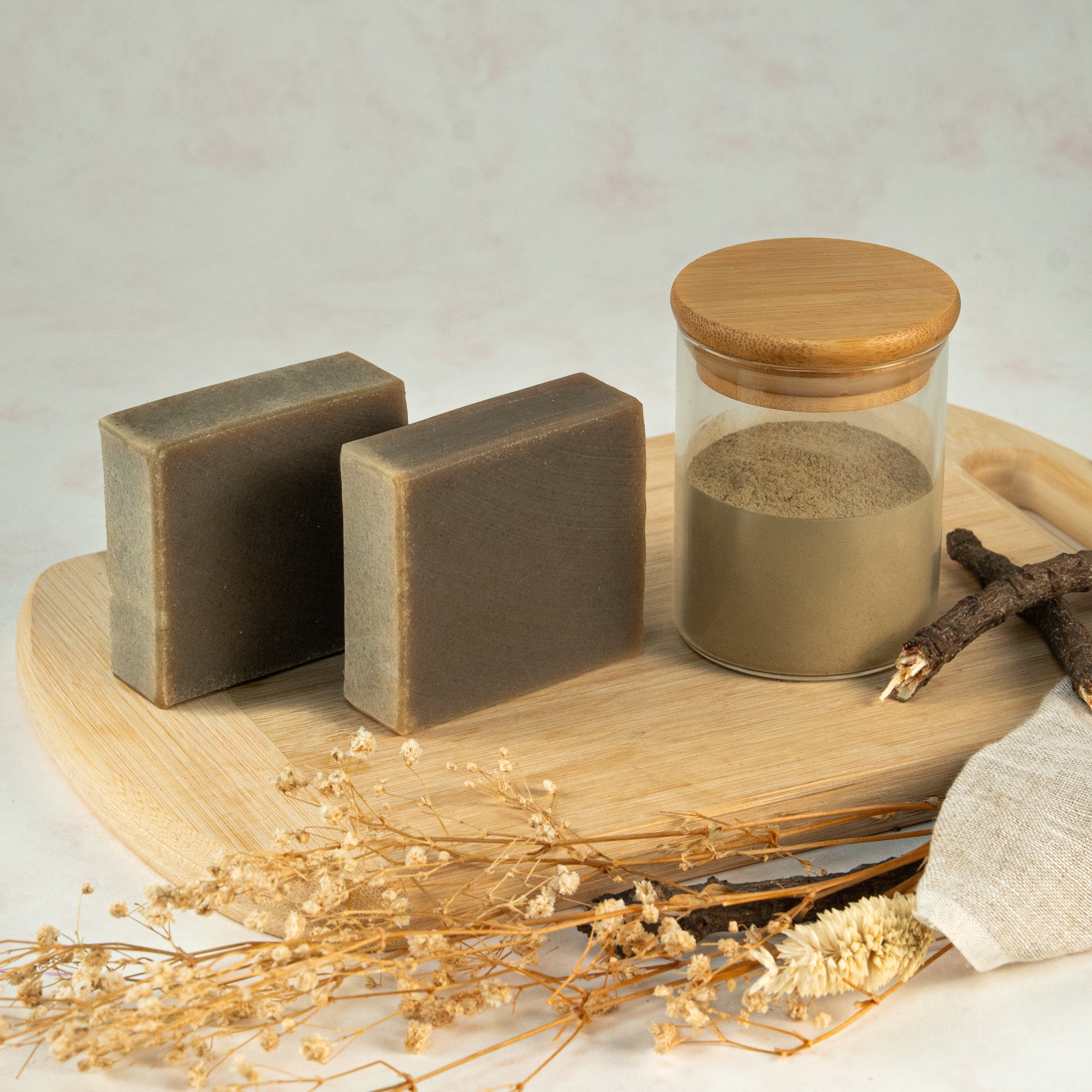 Two bars of soap and a jar with a wooden lid on a wooden tray with dried flowers and a towel.